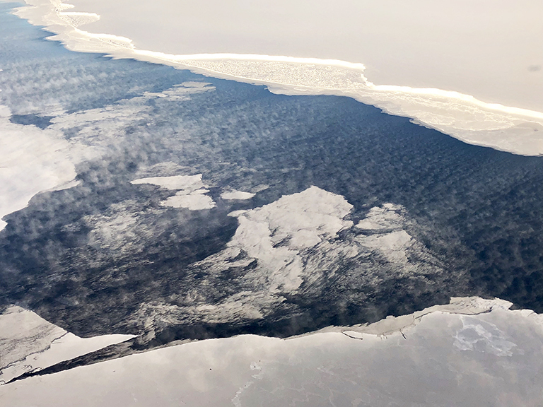 In this photo taken during a NASA IceBridge science flight off of the coast of Antarctica in fall of 2018, dry and cold winds blowing off the iceshelf over an opening in the sea ice (top of figure) cause evaporation from the ocean surface into the atmosphere, where low clouds form. Credit: Linette Boisvert